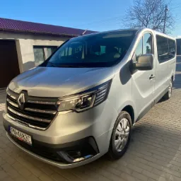 Srebrny Renault Trafic z ciemnymi szybami, zaparkowany na brukowanym podjeździe przed budynkiem z bordowym dachem i brązową bramą garażową, widok z przodu, błękitne niebo.