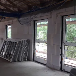 Wnętrze surowego budynku w trakcie montażu okien, widoczne zamontowane okna, okna czekające na montaż oparte o ścianę, siatka zabezpieczająca pod sufitem