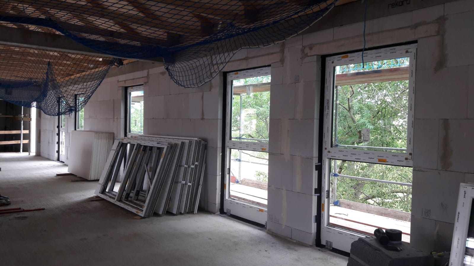 Wnętrze surowego budynku w trakcie montażu okien, widoczne zamontowane okna, okna czekające na montaż oparte o ścianę, siatka zabezpieczająca pod sufitem