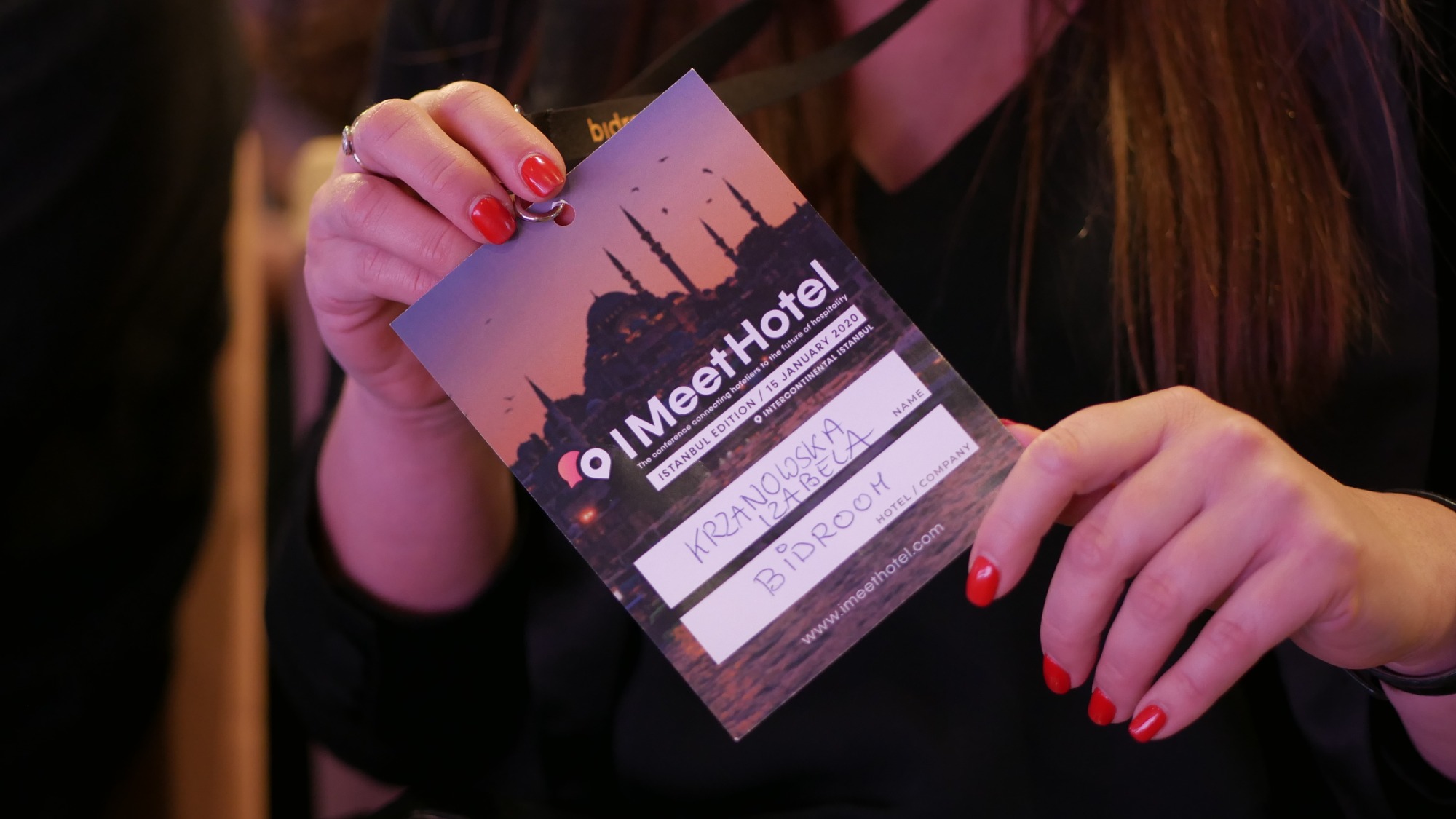 Kobieta trzyma identyfikator konferencji I Meet Hotel z widokiem na Stambuł w tle, z wypełnionymi danymi uczestnika: imię i nazwisko oraz nazwa hotelu.