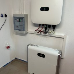 Systemy Oze - Biały falownik Huawei z podłączonymi kablami i biały akumulator energii, zamontowane na białej ścianie w pomieszczeniu technicznym, obok skrzynka z bezpiecznikami.