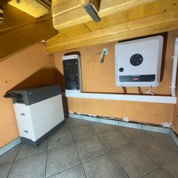 Systemy Oze - Biało-szary moduł magazynu energii, rozdzielnica i falownik zamontowane w narożniku pomieszczenia z pomarańczowymi ścianami i drewnianym stropem.