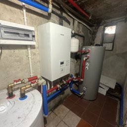 Systemy Oze - Biały piec Panasonic zamontowany w piwnicy, z widocznymi rurami wodnymi w kolorach niebieskim i czerwonym oraz szarym zasobnikiem ciepłej wody, na tle surowych ścian i brudnej podłogi z płytek.