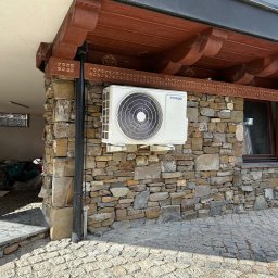 Systemy Oze - Biała jednostka zewnętrzna klimatyzacji marki Hyundai zamontowana na ścianie z kamienia łupanego, pod drewnianym okapem z dekoracyjnymi rzeźbieniami.