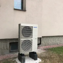 Zewnętrzna jednostka klimatyzacji Panasonic zamontowana na betonowych podstawach z białym kamieniem, przy ścianie budynku z małym oknem.