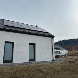 Systemy Oze - Nowoczesny dom jednorodzinny z zainstalowanymi panelami słonecznymi na dachu, widoczny biały van firmy instalacyjnej na posesji, w tle zalesione wzgórza.