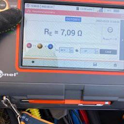 Systemy Oze - Miernik rezystancji uziemienia MPI-540-PV z wynikiem 7,09 Ω, widoczny fragment rękawicy ochronnej i torby narzędziowej firmy Sonel.