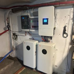 Systemy Oze - Dwa białe magazyny energii Felicitysolar obok ściany z instalacją elektryczną i inwerterem w piwnicy. Widoczne rury i dywan na podłodze.