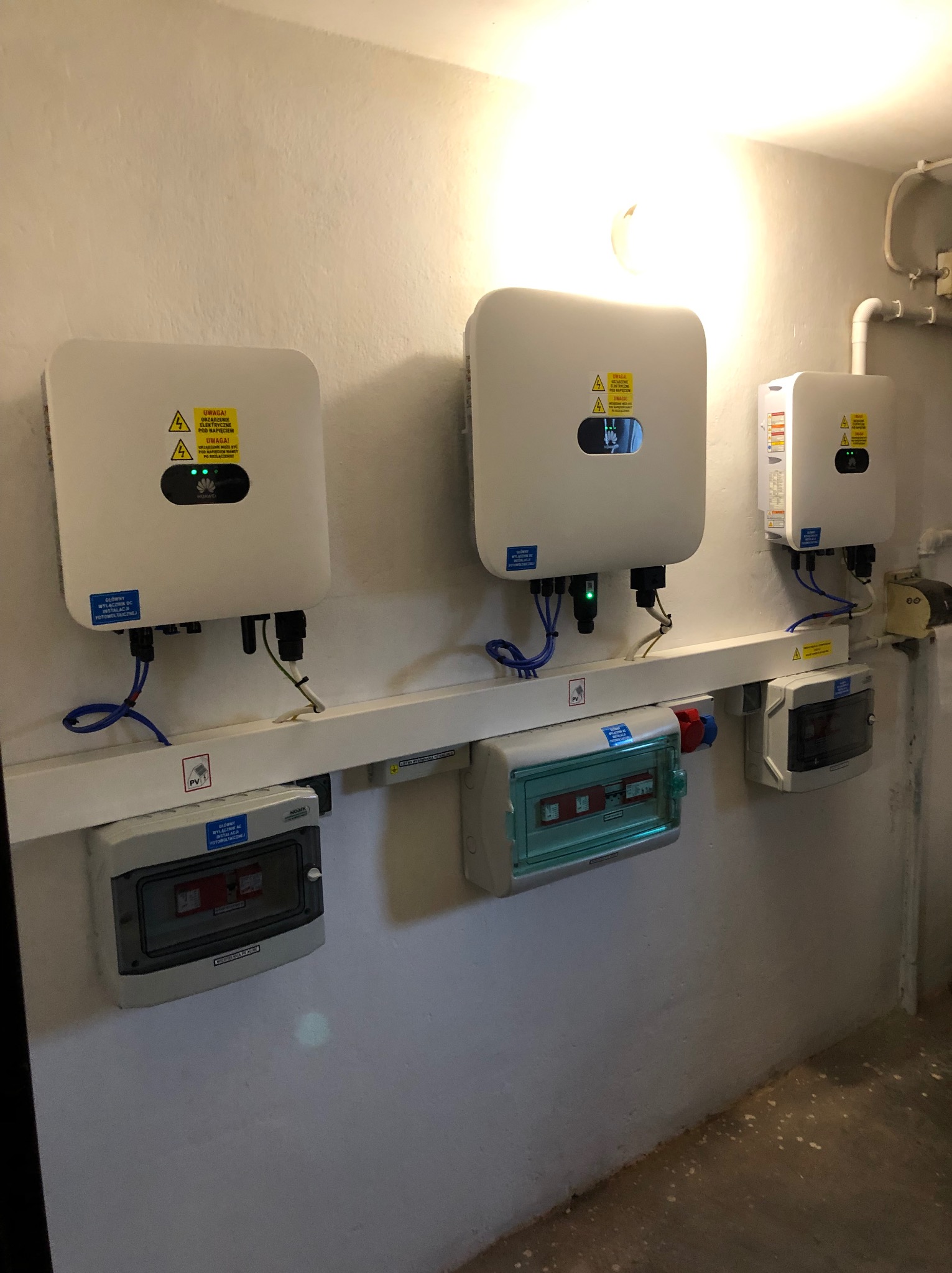 Trzy falowniki Huawei zamontowane na ścianie obok skrzynek bezpiecznikowych. Widoczne przewody i oznaczenia ostrzegawcze.