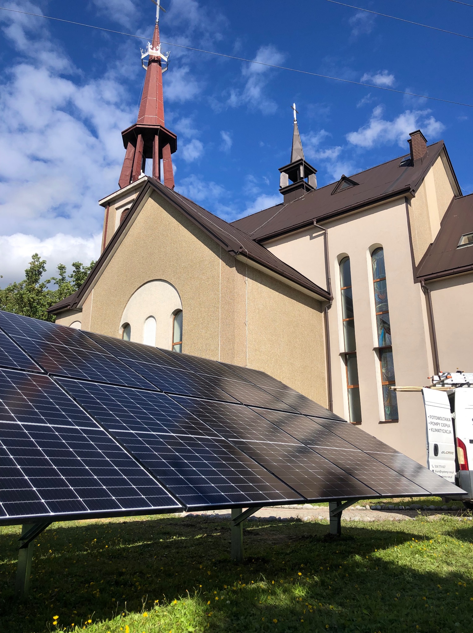 Instalacja paneli słonecznych na trawniku przed kościołem z czerwoną wieżą, widoczny biały bus z reklamą usług.