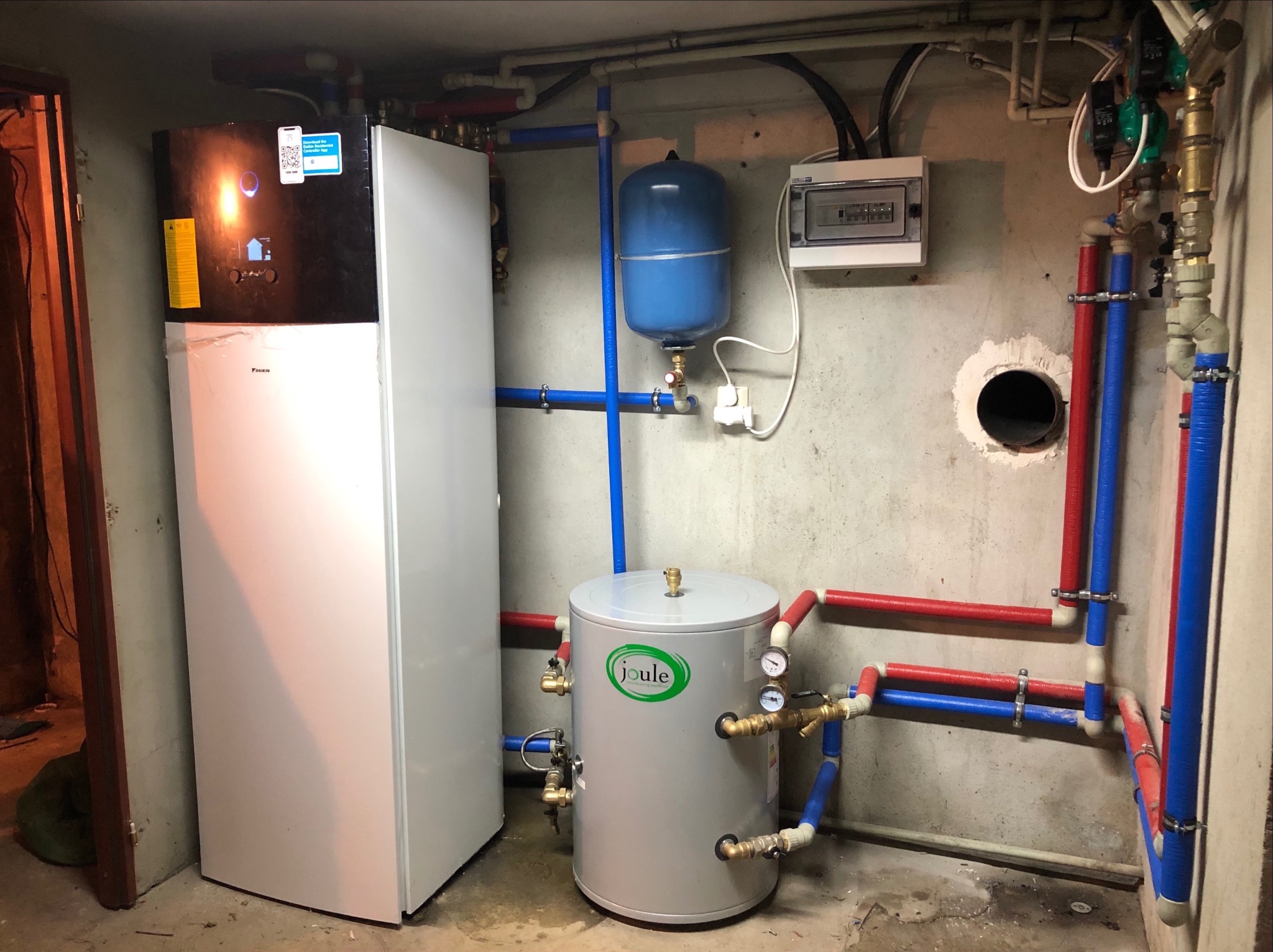 Zainstalowana pompa ciepła Daikin z zasobnikiem Joule, widoczne rury instalacyjne w kolorach niebieskim i czerwonym oraz osprzęt elektryczny na ścianie w surowym pomieszczeniu.