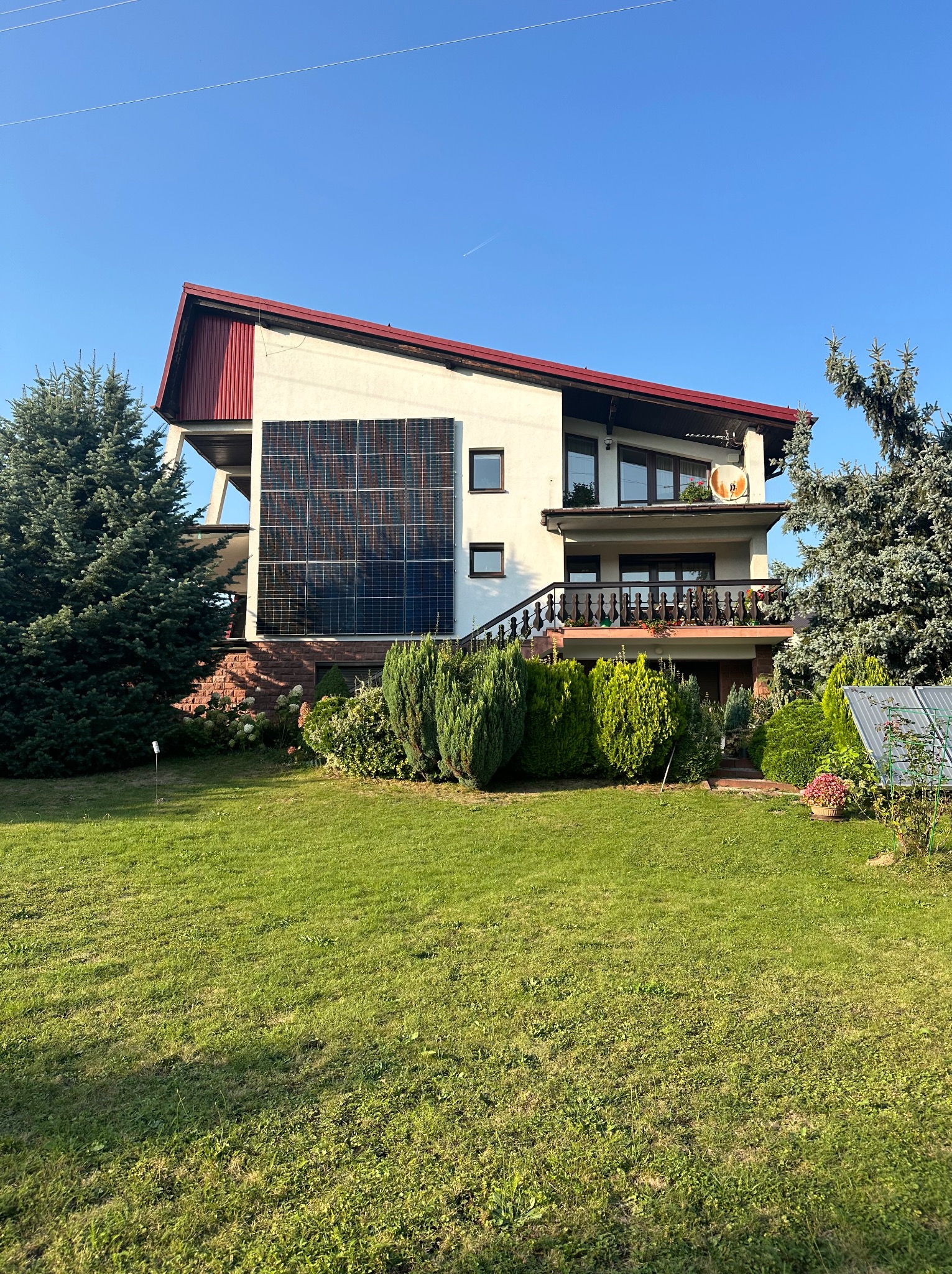 Dom jednorodzinny z panelami słonecznymi na ścianie, otoczony zielonym trawnikiem i drzewami iglastymi, widok z przodu w słoneczny dzień.