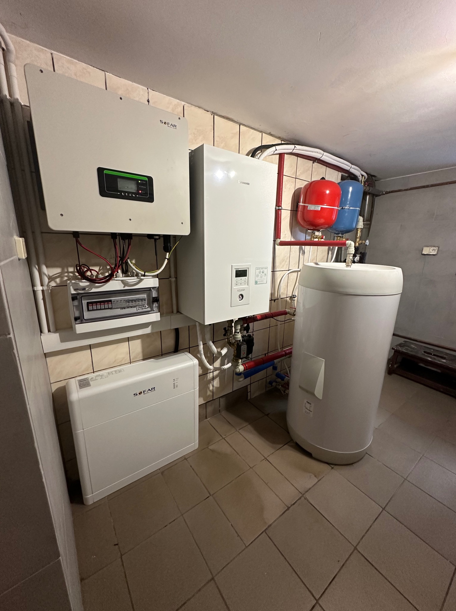 Kotłownia z zainstalowanym inwerterem Sofar, rozdzielnicą elektryczną, bojlerem i zbiornikami wyrównawczymi, całość na tle ceramicznych płytek ściennych i podłogowych.