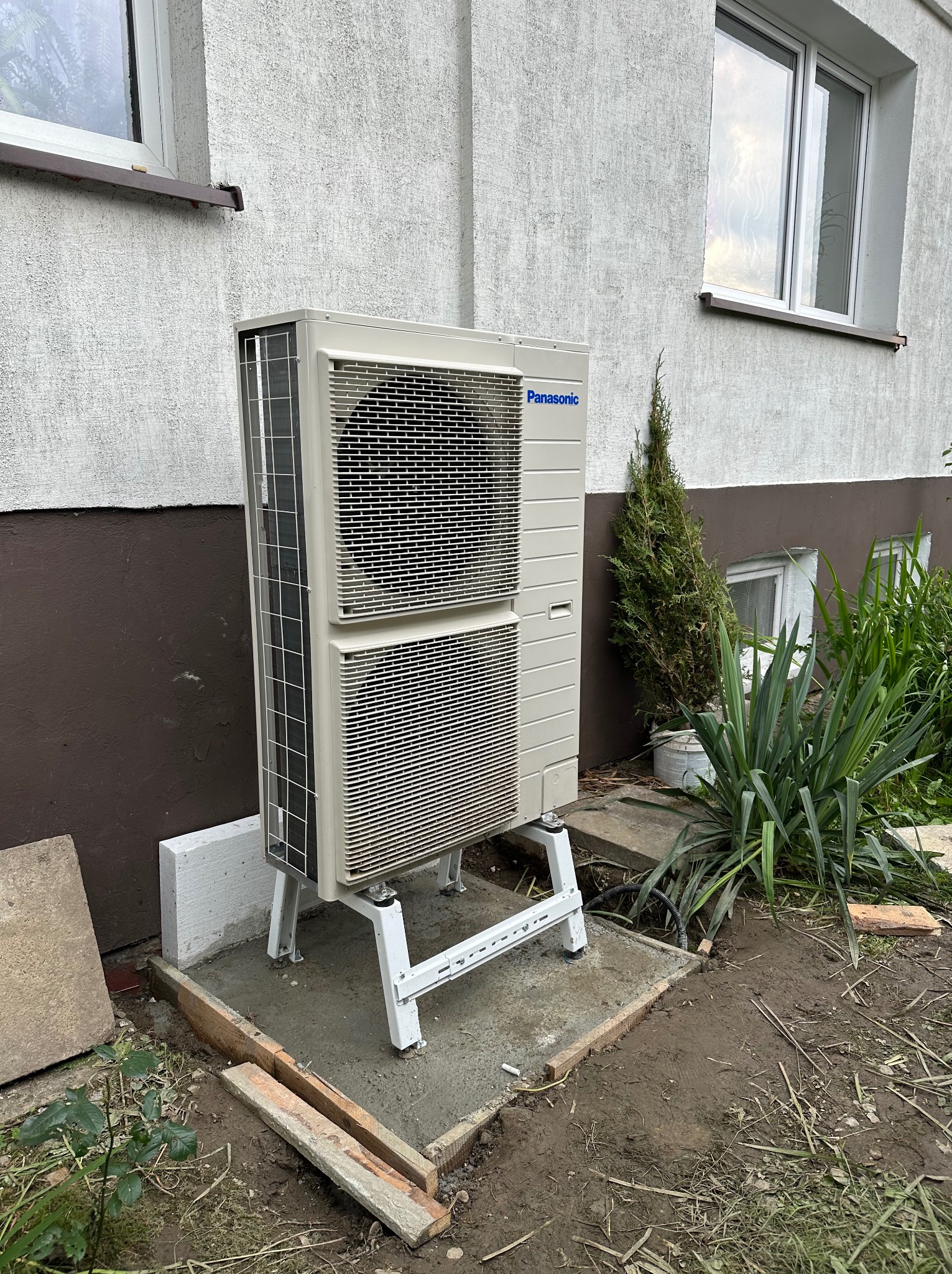Zewnętrzna jednostka pompy ciepła Panasonic zamontowana na betonowej podstawie z białym stelażem, widoczna z boku budynku z oknami i roślinnością.