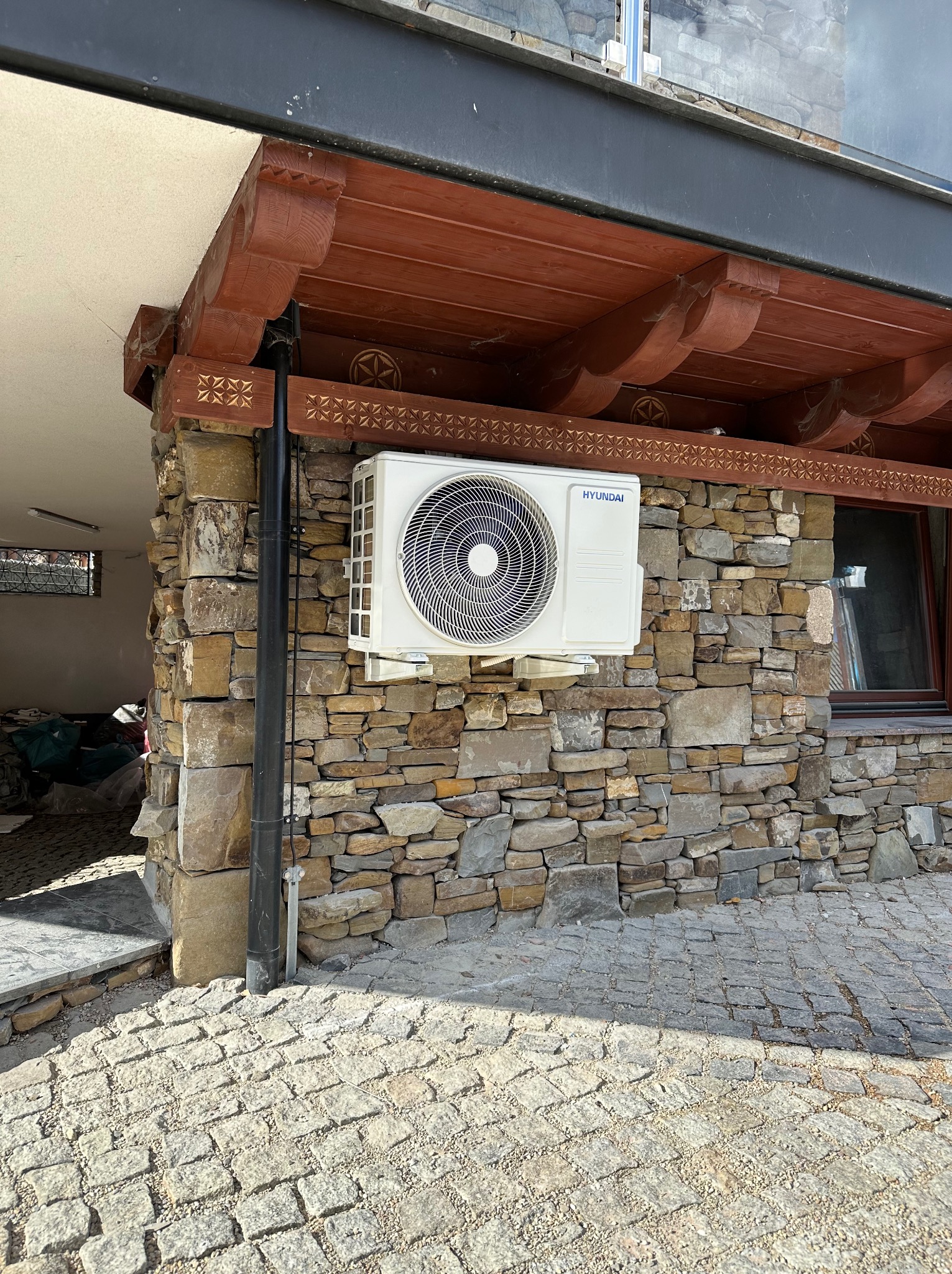 Biała jednostka zewnętrzna klimatyzacji marki Hyundai zamontowana na ścianie z kamienia łupanego, pod drewnianym okapem z dekoracyjnymi rzeźbieniami.