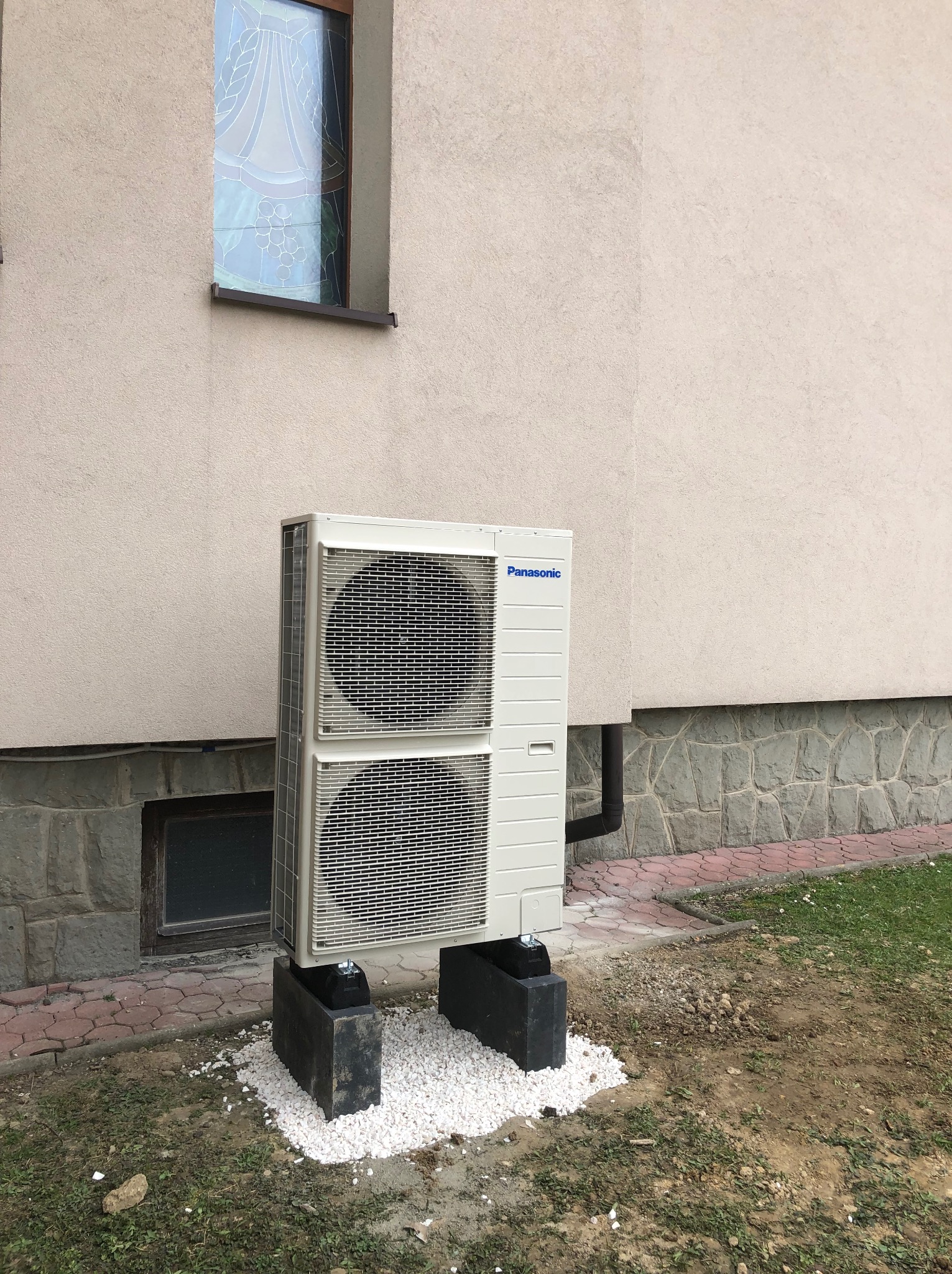 Zewnętrzna jednostka klimatyzacji Panasonic zamontowana na betonowych podstawach z białym kamieniem, przy ścianie budynku z małym oknem.