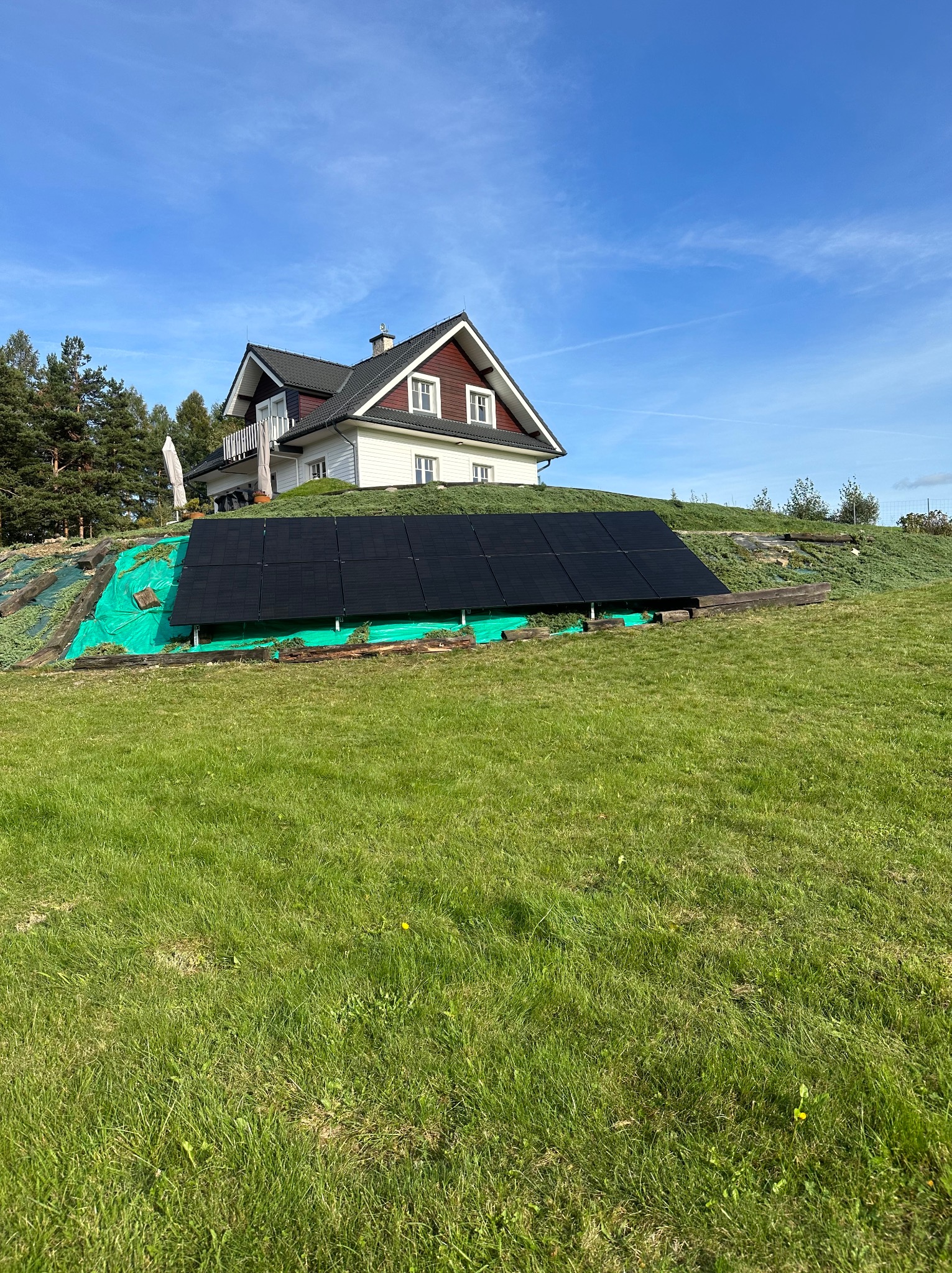 Czarny panel słoneczny zainstalowany na zielonym zboczu wzgórza, w tle biały dom z czerwonymi akcentami i błękitne niebo.