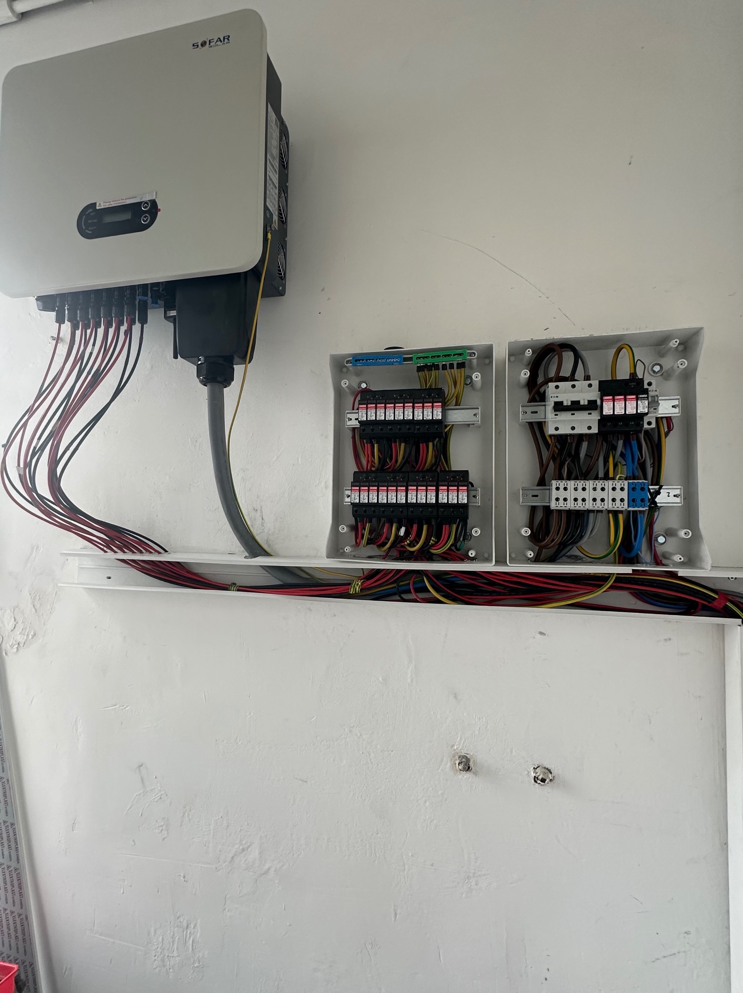 Biały inwerter SOFAR przymocowany do ściany z dwoma otwartymi skrzynkami elektrycznymi poniżej, eksponującymi wewnętrzne okablowanie i zabezpieczenia; widoczne kable prowadzące do inwertera...