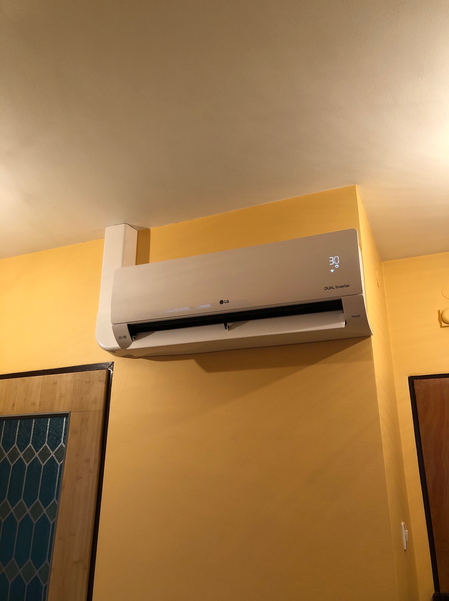 Klimatyzator LG Dual Inverter zamontowany na żółtej ścianie w pomieszczeniu, widoczny wyświetlacz z temperaturą 30 stopni Celsjusza.