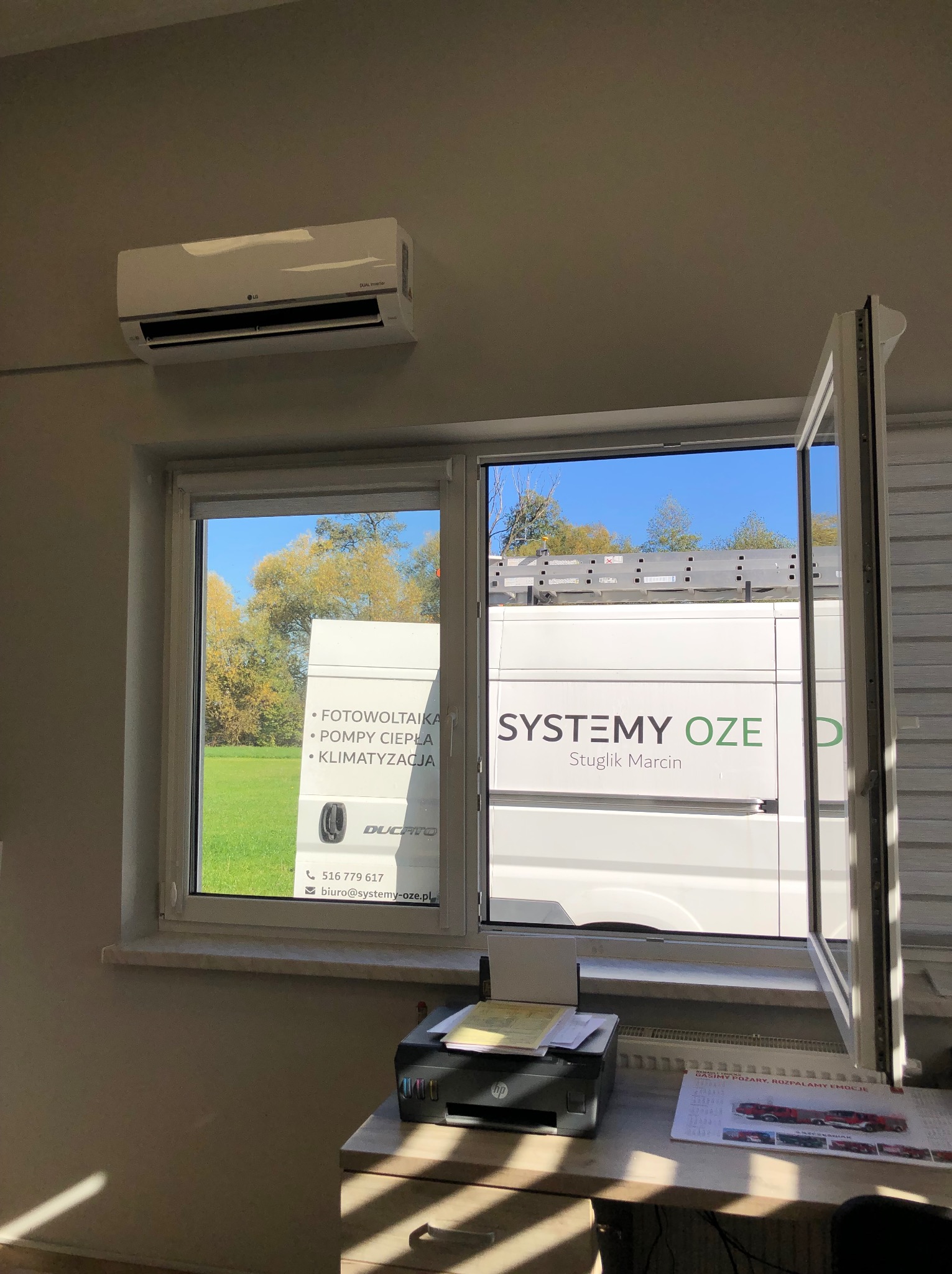 Wnętrze biura z widokiem przez okno na zaparkowany samochód dostawczy firmy instalującej odnawialne źródła energii, na ścianie klimatyzator LG.