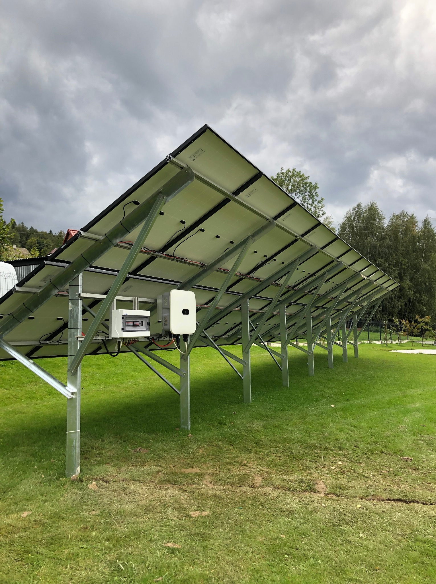 Widok od spodu na instalację paneli słonecznych na metalowej konstrukcji wspartej na słupkach wbitych w trawnik, z widoczną białą skrzynką rozdzielczą i inwerterem.