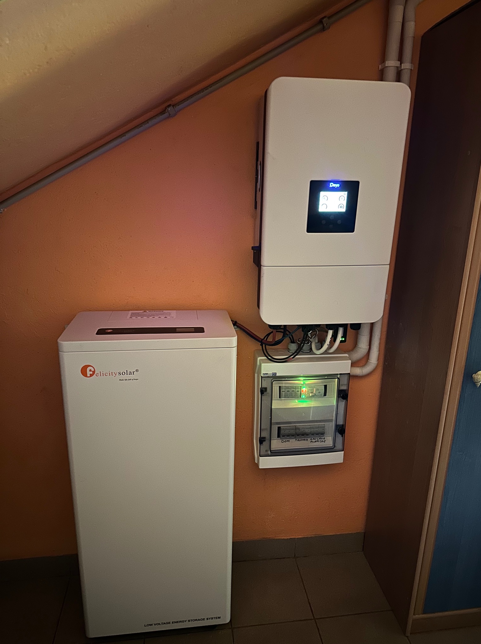 Biały magazyn energii Felicity Solar obok falownika Deye i skrzynki z bezpiecznikami na pomarańczowej ścianie. Widoczne przewody i rury instalacyjne.