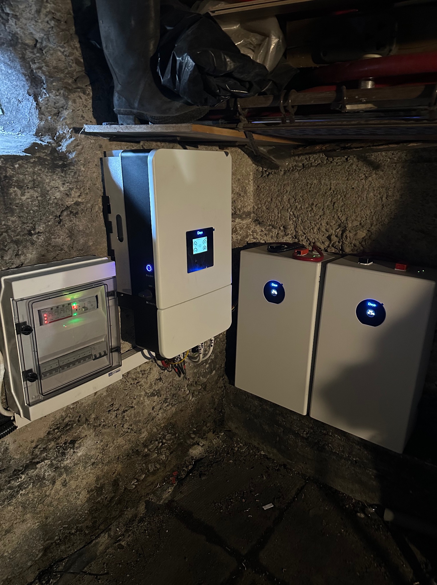 Instalacja magazynu energii w piwnicy o kamiennych ścianach. Widoczna skrzynka elektryczna, inwerter i dwa akumulatory. Elementy połączone przewodami.