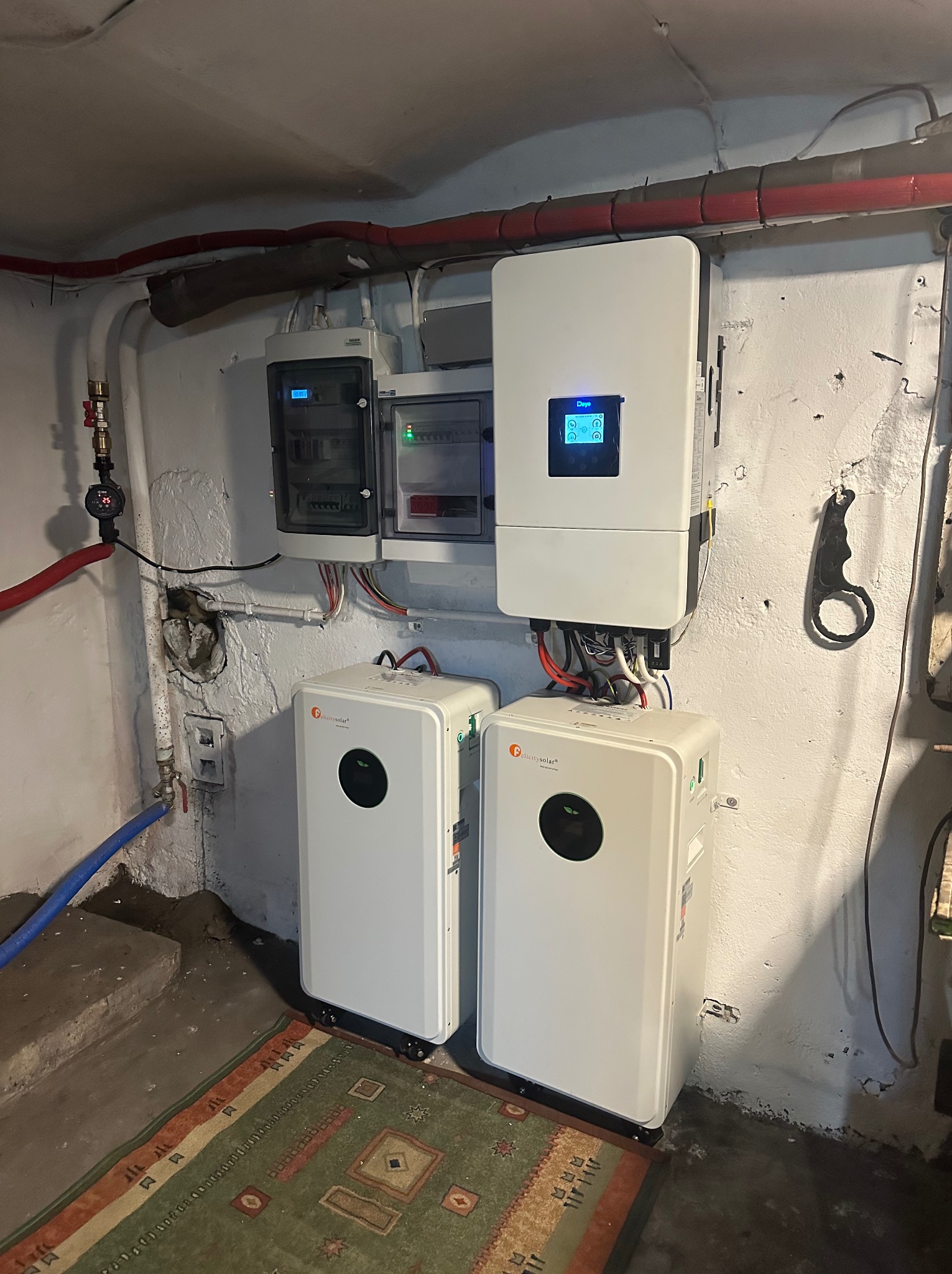 Dwa białe magazyny energii Felicitysolar obok ściany z instalacją elektryczną i inwerterem w piwnicy. Widoczne rury i dywan na podłodze.