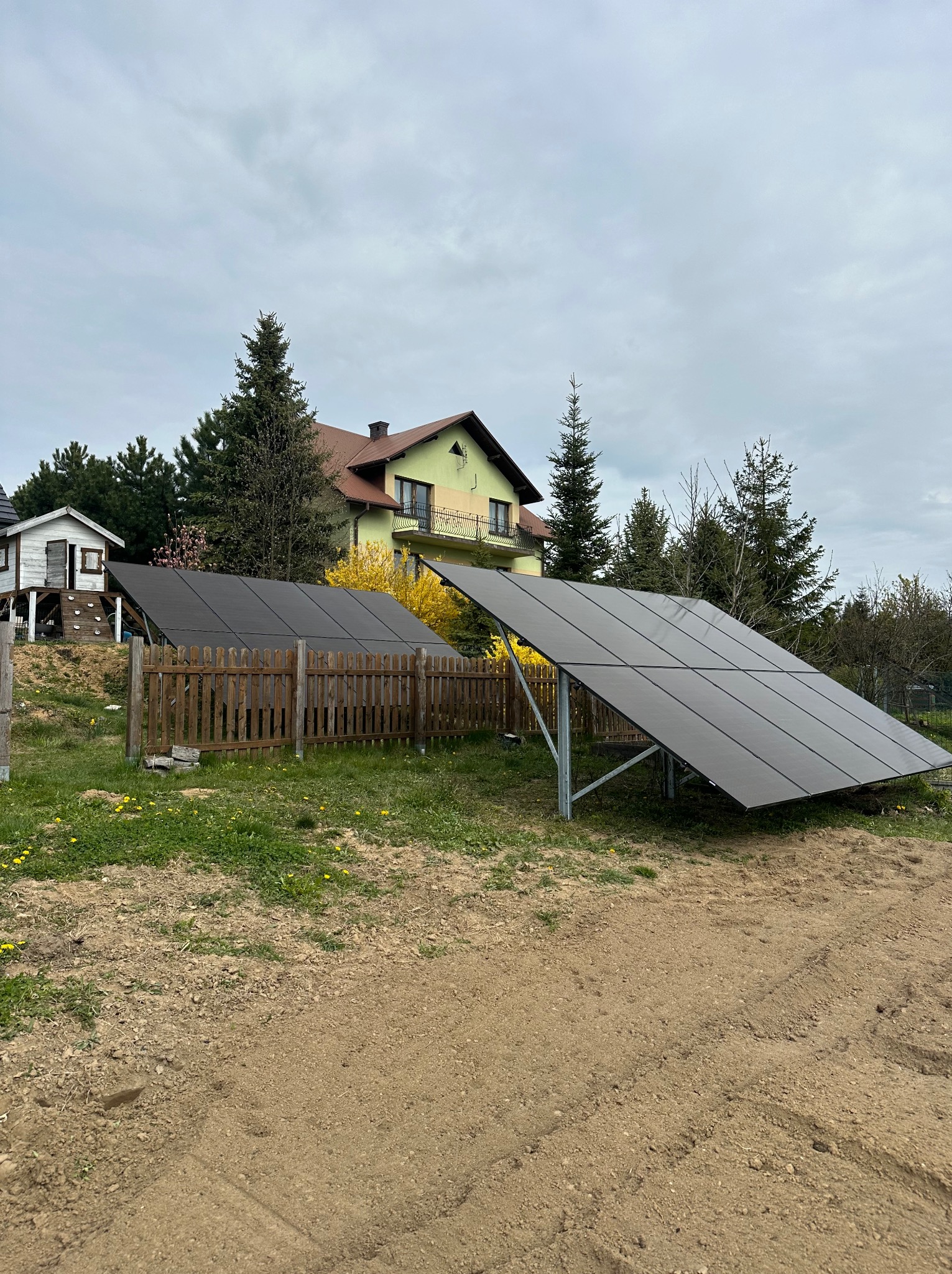 Panele fotowoltaiczne na gruncie, za drewnianym płotem, z domem w tle. Instalacja w trakcie realizacji, widoczne elementy montażowe i przygotowany teren.