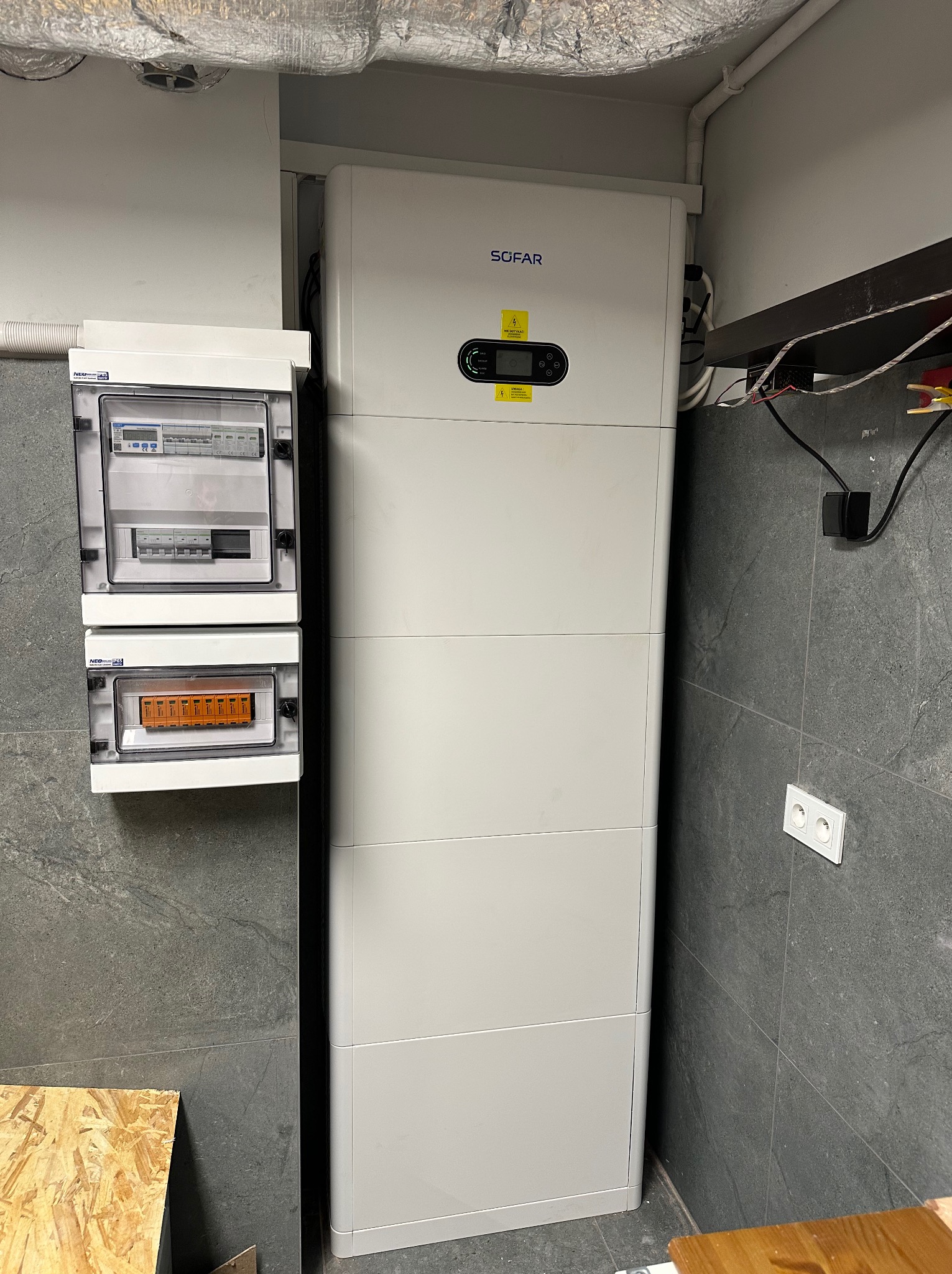 Biały modułowy magazyn energii Sofar obok skrzynki elektrycznej na szarej ścianie z kamienia. Widoczne przewody i gniazdko elektryczne. Instalacja w pomieszczeniu.