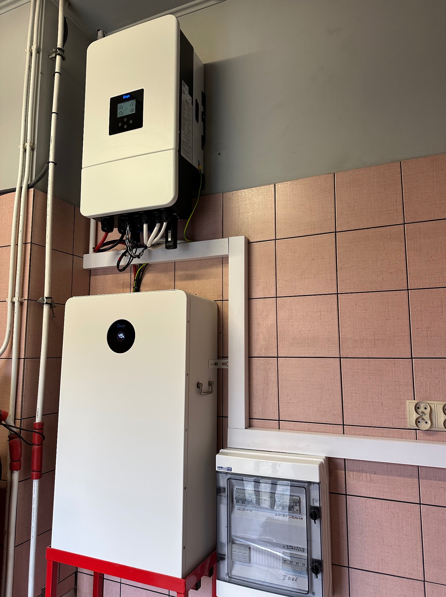 Biały inwerter i magazyn energii Deye zamontowane na ścianie z różowych płytek, widoczna skrzynka elektryczna. Instalacja w pomieszczeniu technicznym.