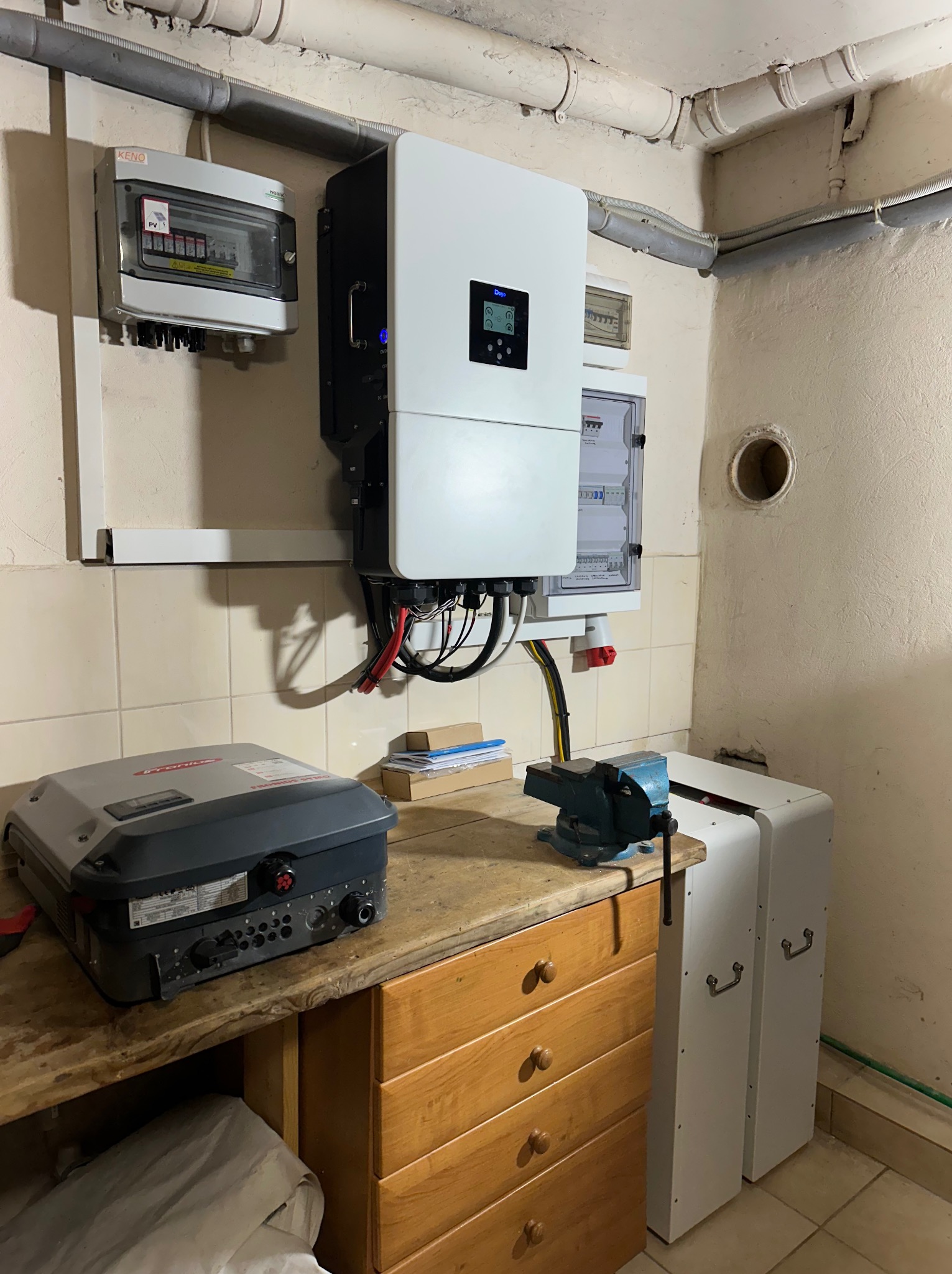 Zamontowany inwerter fotowoltaiczny Fronius, rozdzielnica i magazyn energii w piwnicy. Widoczne rury instalacyjne pod sufitem i płytki na ścianach.