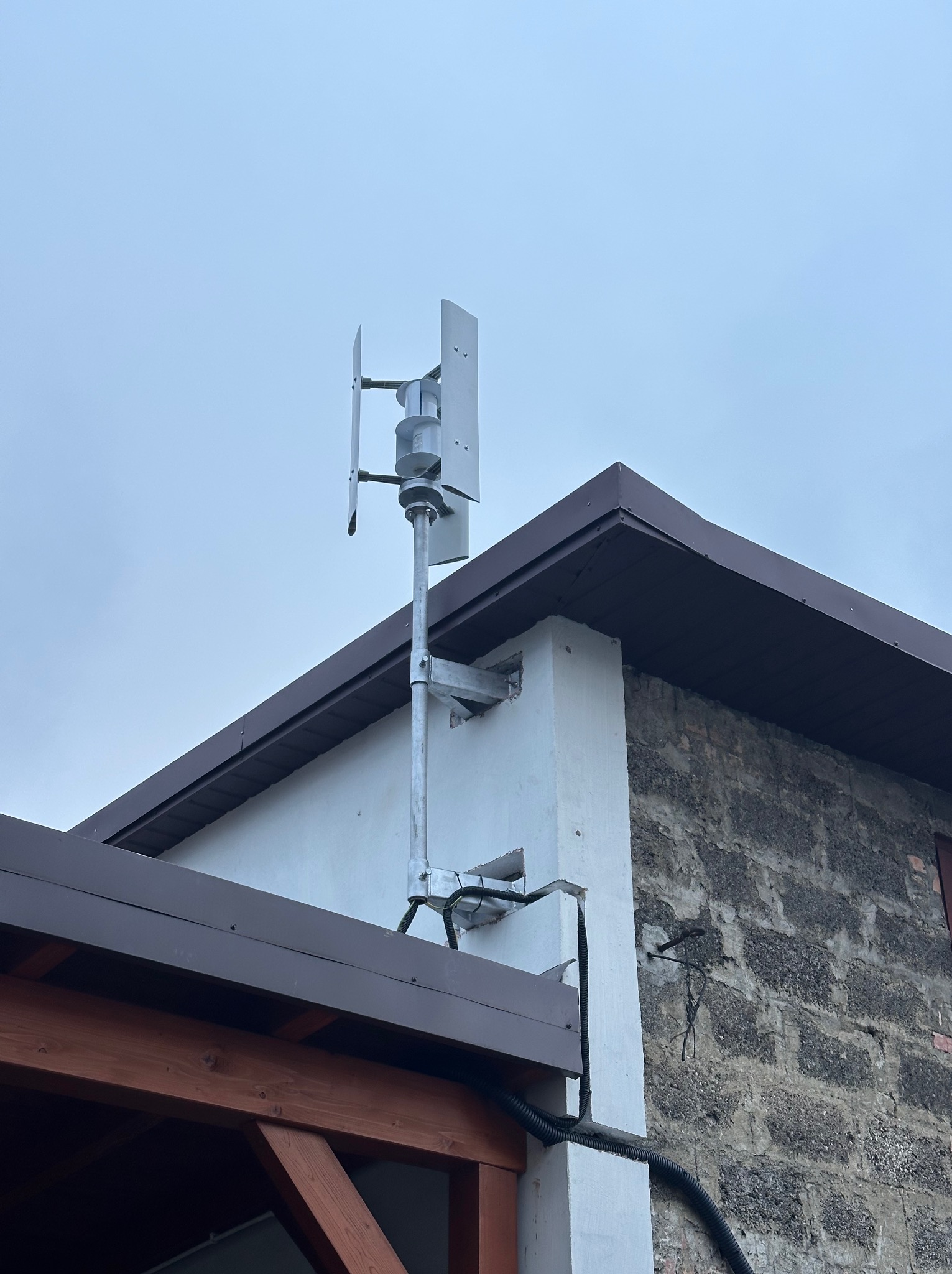 Antena na elewacji budynku z brązowym dachem, zamontowana na metalowym stelażu. Widoczne przewody i fragment drewnianej konstrukcji. Pochmurne niebo w tle.