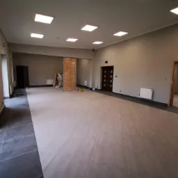 Duże, puste pomieszczenie biurowe z jasnymi płytkami podłogowymi, grzejnikami na ścianach i drzwiami zabezpieczonymi folią ochronną, przygotowane do dalszych prac wykończeniowych.