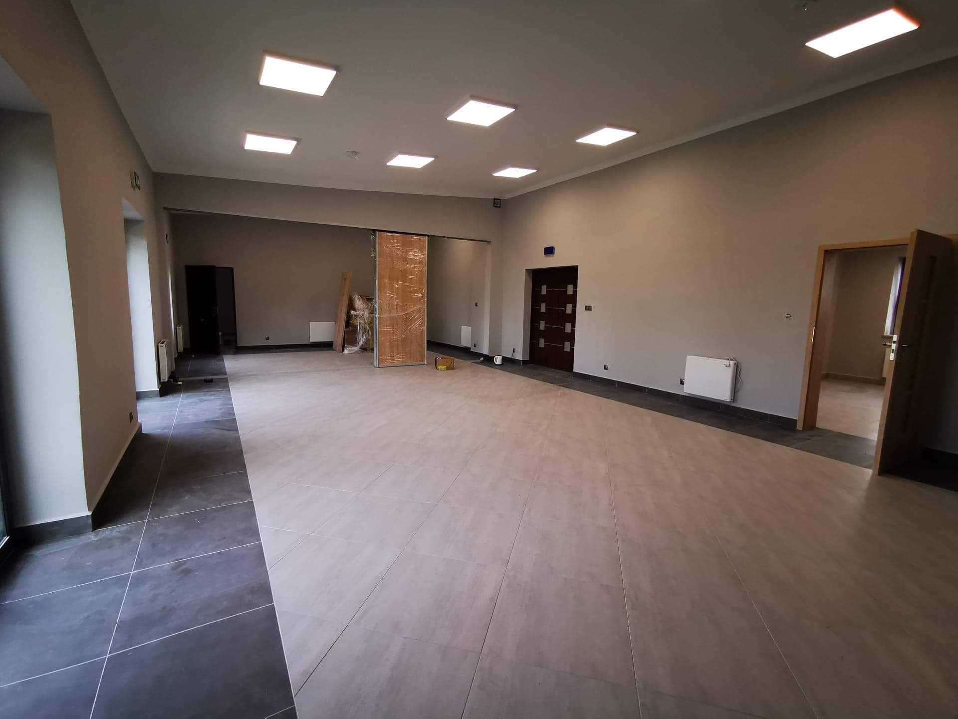 Duże, puste pomieszczenie biurowe z jasnymi płytkami podłogowymi, grzejnikami na ścianach i drzwiami zabezpieczonymi folią ochronną, przygotowane do dalszych prac wykończeniowych.