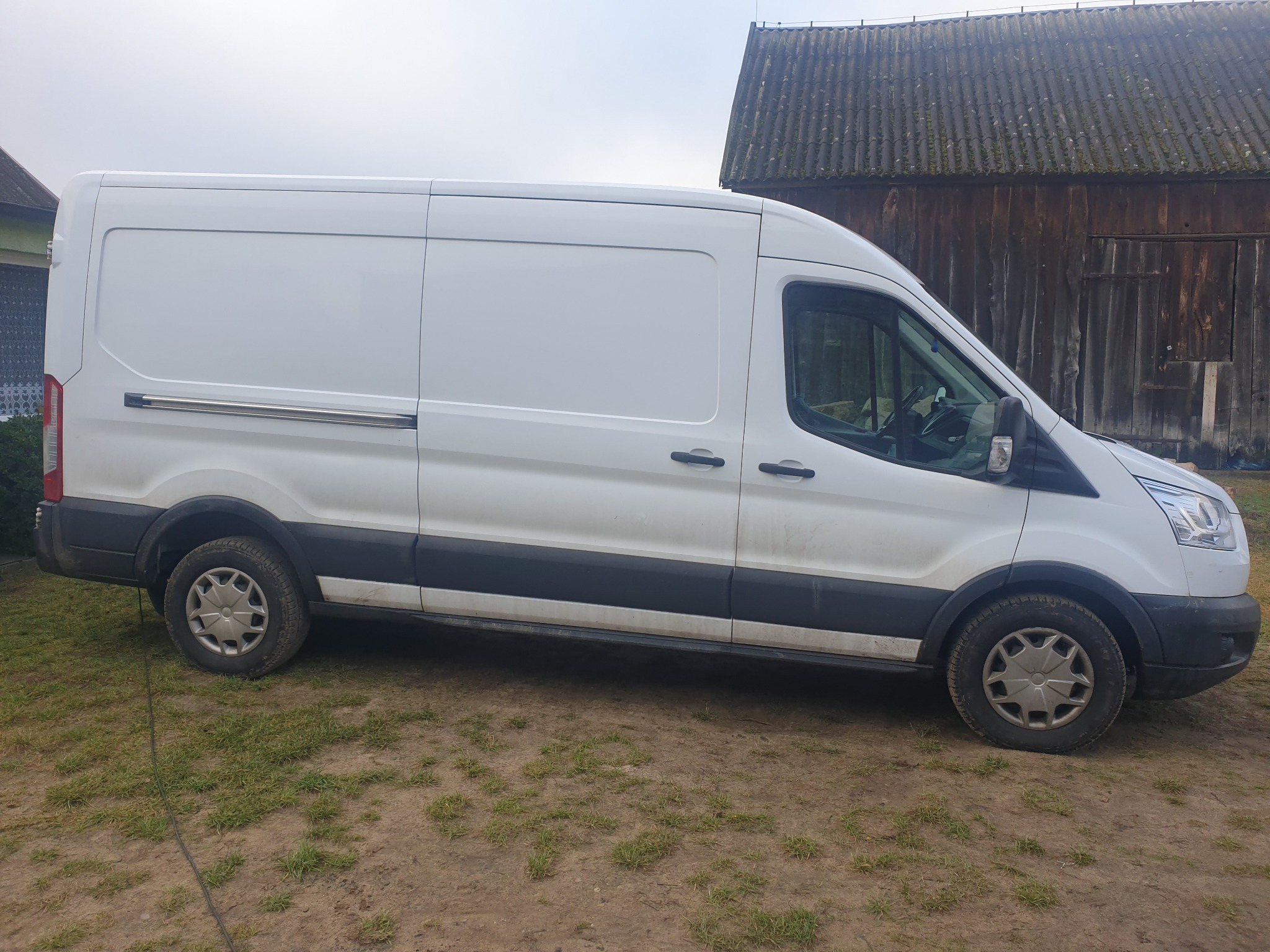 Biały samochód dostawczy Ford Transit zaparkowany na trawiastym terenie przed drewnianym budynkiem gospodarczym z zadaszeniem pokrytym mchem.