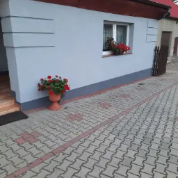 Jasnoszara elewacja domu jednorodzinnego z czerwoną dachówką, ozdobiona donicami z czerwonymi kwiatami przy oknie i obok wejścia, widoczna kostka brukowa na podwórku.