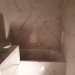 Wykończona łazienka z wanną obudowaną białymi płytkami imitującymi marmur i umywalką nablatową, widoczne narzędzia hydrauliczne na podłodze.