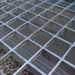 Metalowa krata pomostowa z widoczną strukturą kwadratową, lekko zniekształconą perspektywą i drobnym żwirem w tle.