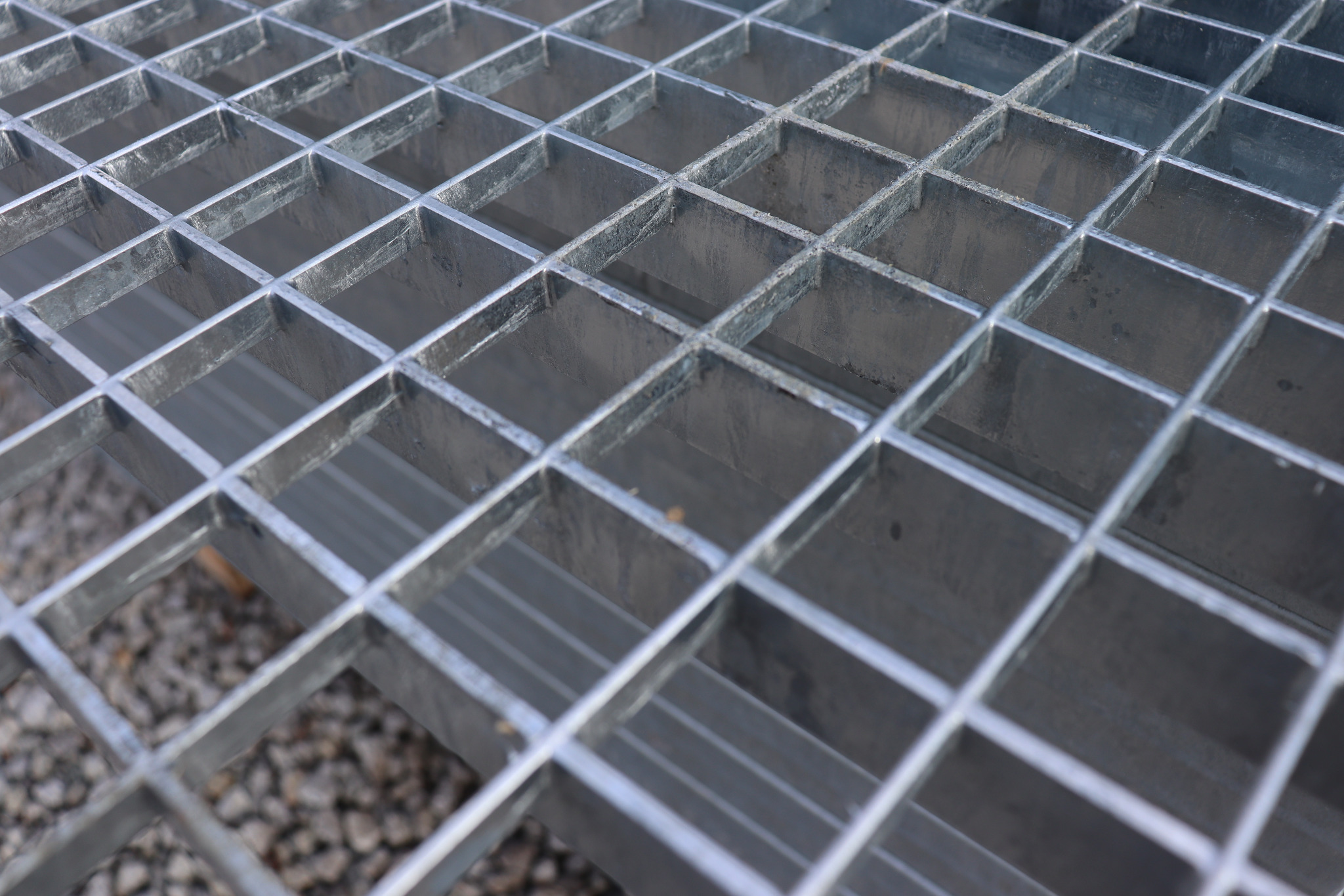 Metalowa krata pomostowa z widoczną strukturą kwadratową, lekko zniekształconą perspektywą i drobnym żwirem w tle.
