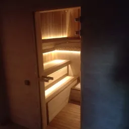 Wykończona sauna z drewnianymi ławkami i podświetleniem LED, widoczna z zewnątrz przez otwarte drzwi z drewnianą klamką.