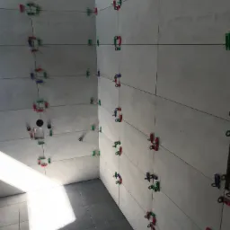 Świeżo ułożone, szare płytki na ścianach i podłodze łazienki, z widocznymi kolorowymi klipsami poziomującymi i czarnymi liniami fug, w trakcie prac wykończeniowych.