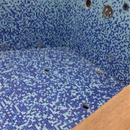 Wnętrze wanny jacuzzi wyłożonej mozaiką z niebieskich i białych płytek, widoczne otwory na dysze i odpływ, drewniane wykończenie krawędzi.