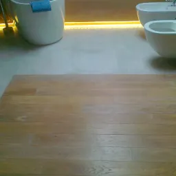 Jasna, drewniana podłoga w łazience, z widoczną wanną i toaletą w tle, oświetlona subtelnym, ciepłym światłem LED pod wanną.