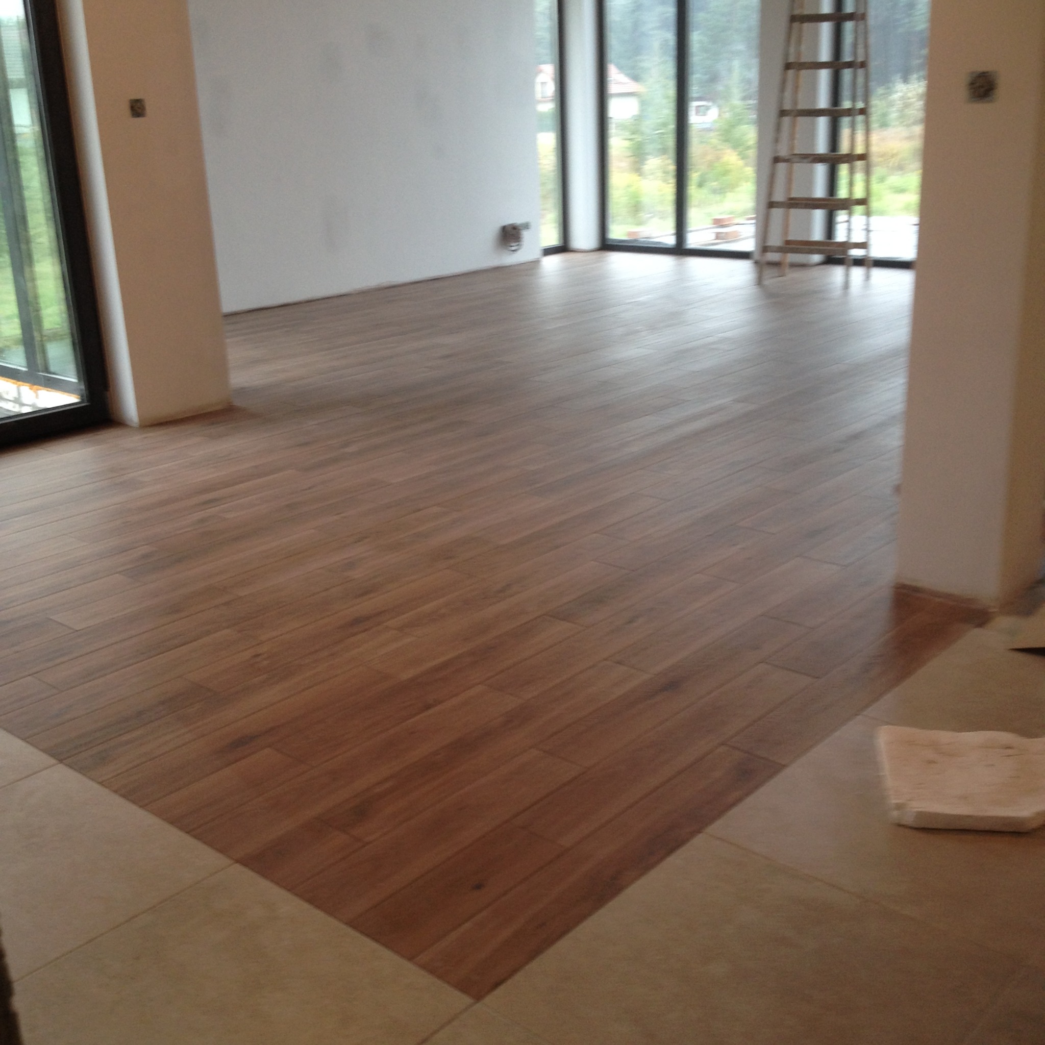 Ułożona podłoga z paneli imitujących drewno, widoczne fragmenty białych ścian i okna z widokiem na zewnątrz, drabina oparta o ścianę w tle