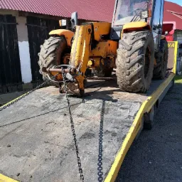 Żółta ładowarka teleskopowa zabezpieczona łańcuchami na żółtej lawecie, widok z przodu na tle budynku z czerwonym dachem.