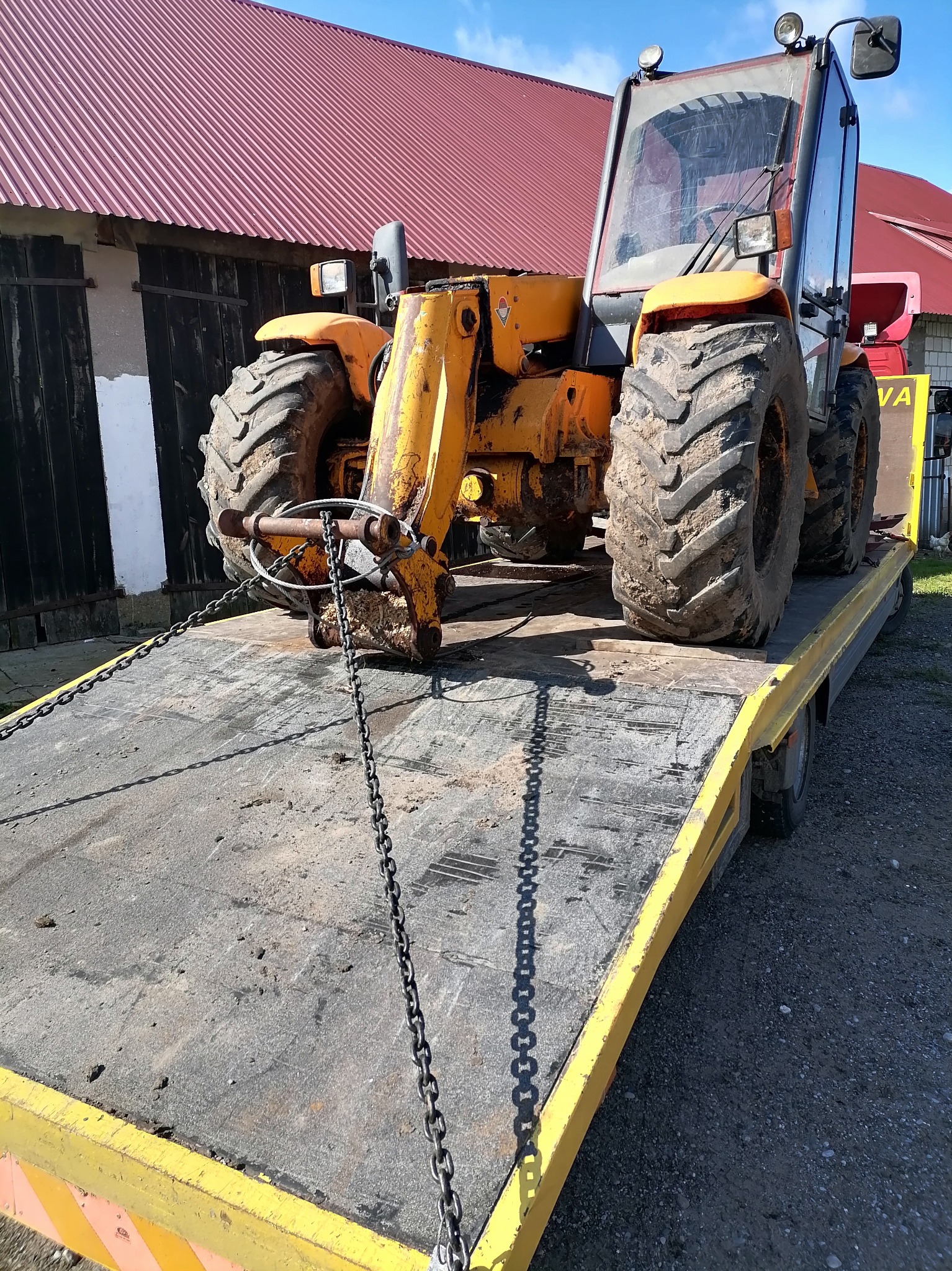 Żółta ładowarka teleskopowa zabezpieczona łańcuchami na żółtej lawecie, widok z przodu na tle budynku z czerwonym dachem.
