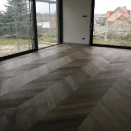 Ułożony parkiet w jodełkę w nowym domu z dużymi oknami, widok na ogród i budynek w tle.