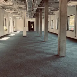 Przestronne, jasne wnętrze biurowe z szarą wykładziną w kratę, betonowymi filarami i widocznymi instalacjami pod sufitem.