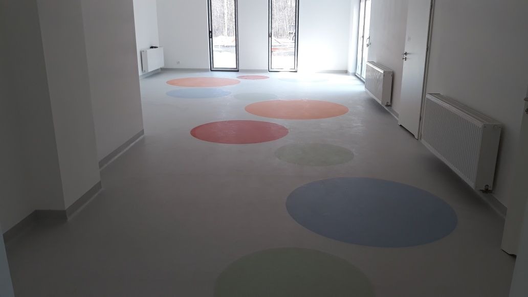 Wnętrze z szarą podłogą w kolorowe, okrągłe wzory, widoczne białe ściany z grzejnikami i drzwiami, oraz okna wychodzące na zimowy krajobraz.